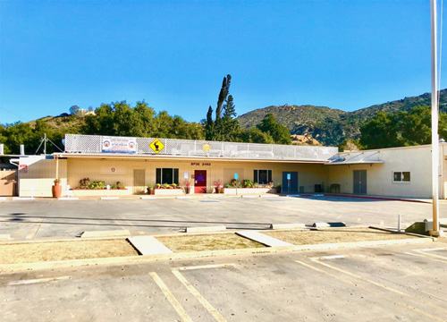 A single-story beige building with colorful doors and a large banner featuring various logos. The building is set against a backdrop of hills and clear blue sky, with an empty parking lot in the foreground.
