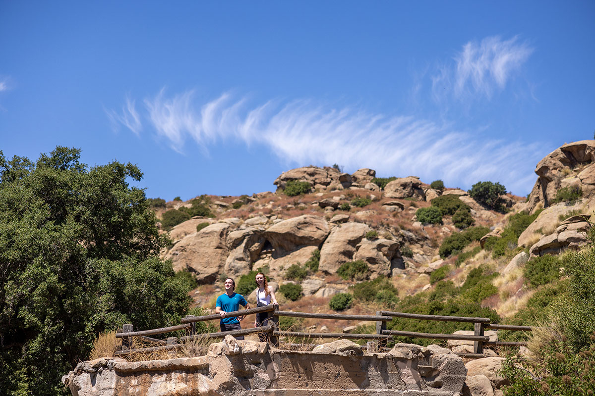 Two people stand on a rocky platform surrounded by trees and large boulders under a blue sky with wispy clouds.